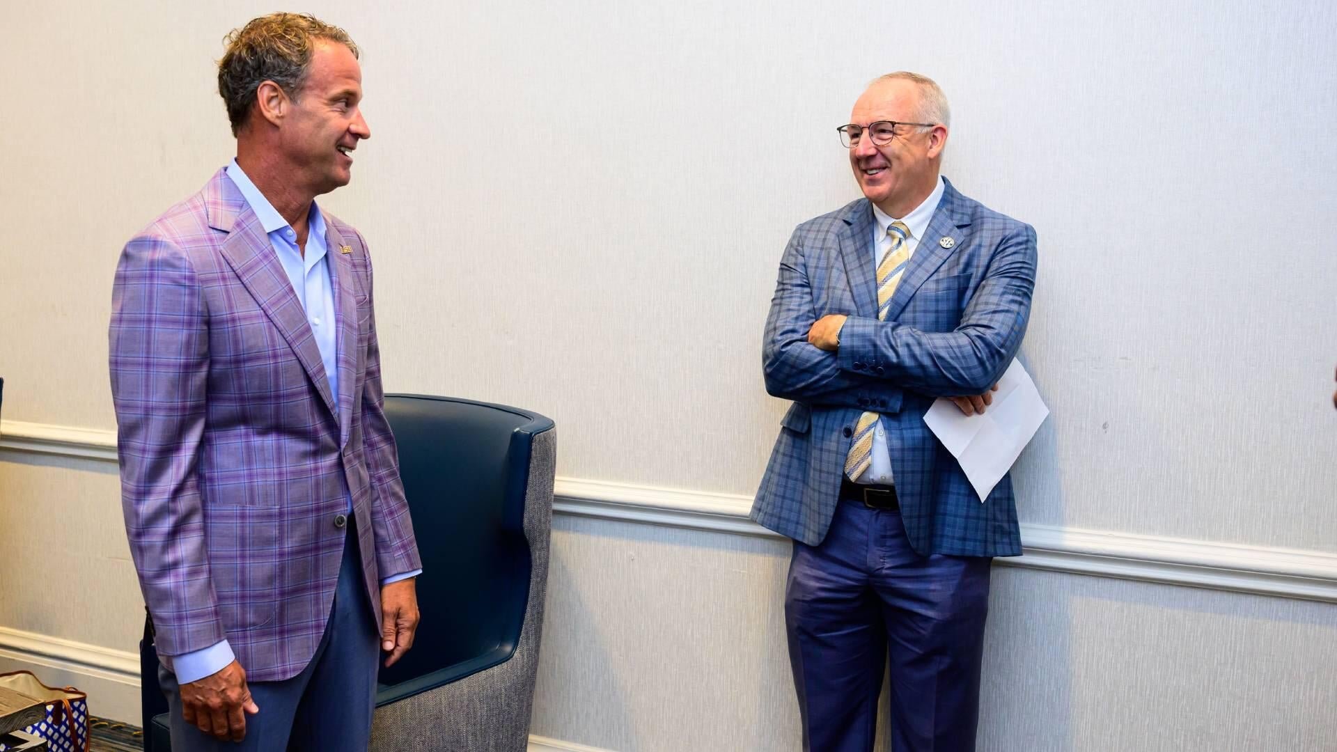 Two men in plaid blazers smile while conversing in a light-colored room, one holding papers.