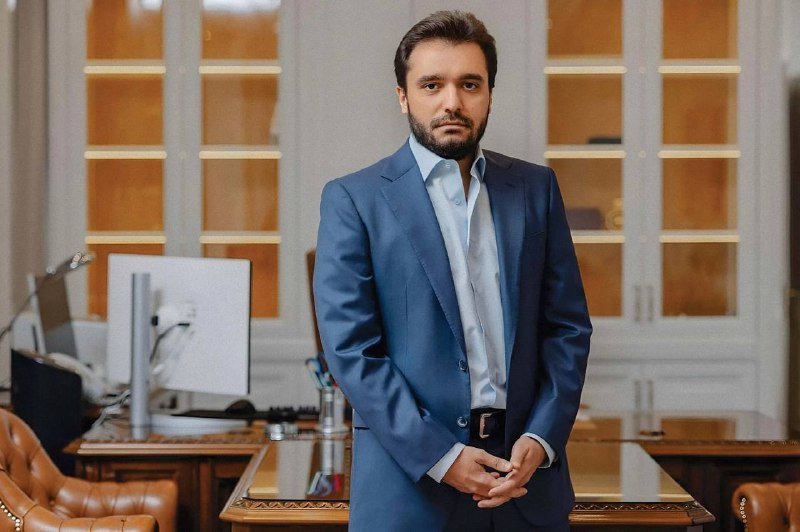 A bearded man in a blue suit and light blue shirt stands with hands clasped in an office featuring a computer monitor, desk, and glass-paned wooden cabinets.