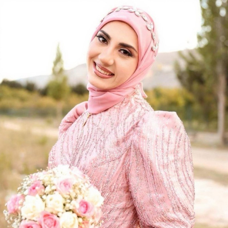 A smiling woman wearing a pink beaded hijab and matching embellished dress holds a bouquet of pink and white roses outdoors.