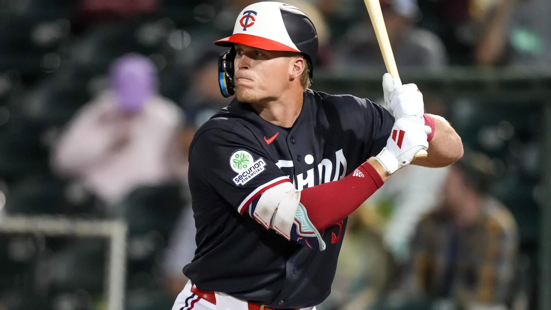 A baseball player in a navy Minnesota Twins jersey and helmet swings a wooden bat during a game.