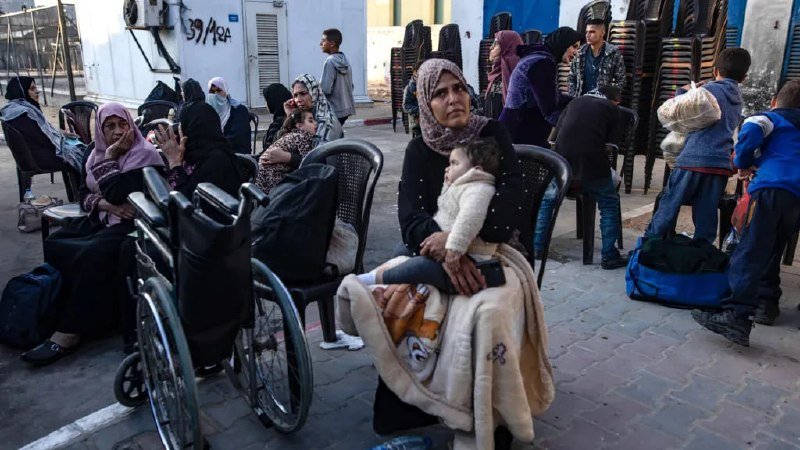 A woman in a headscarf holds a child wrapped in a blanket while seated on a plastic chair, surrounded by other women, children, and stacked chairs on a sidewalk near a building.