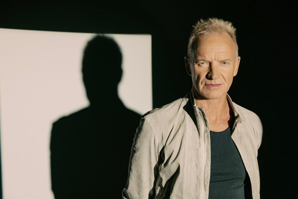 A man with short blonde hair wearing a light jacket over a dark shirt stands against a black backdrop, with his shadow cast on a lit panel behind him.
