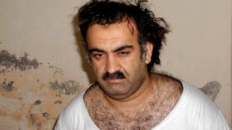 A disheveled man with dark curly hair and a mustache wearing a white t-shirt stands against a stained, weathered wall.