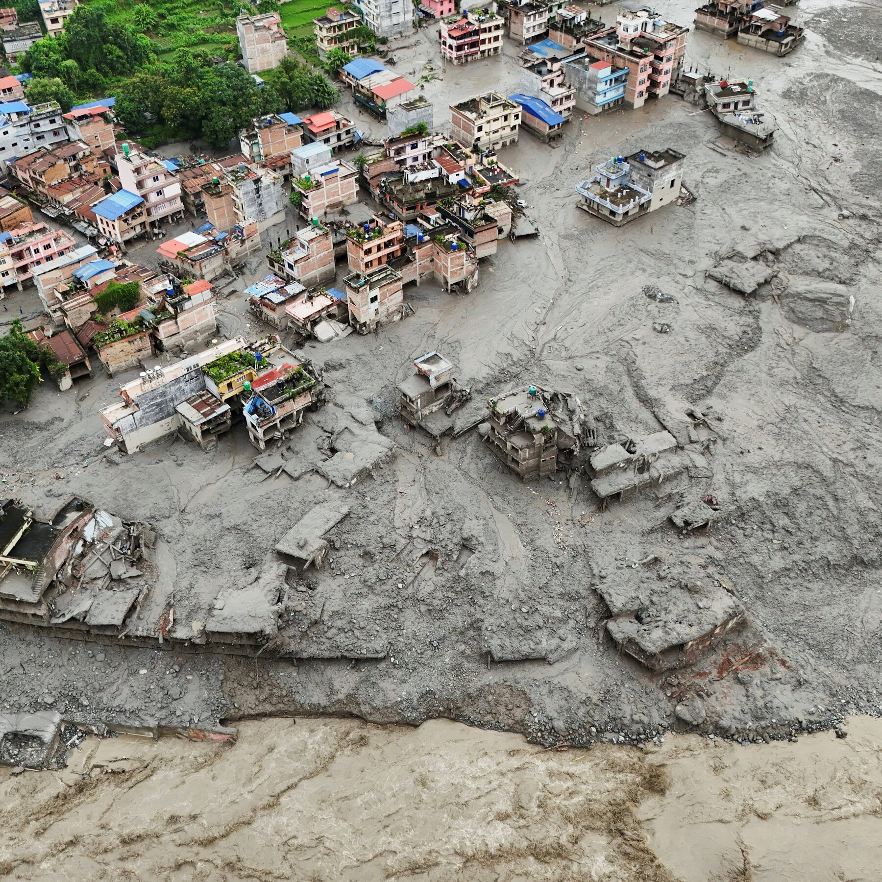Aerial view shows a densely packed town partially buried under thick gray mud and debris, with a rushing muddy river flowing along the bottom edge of the frame.