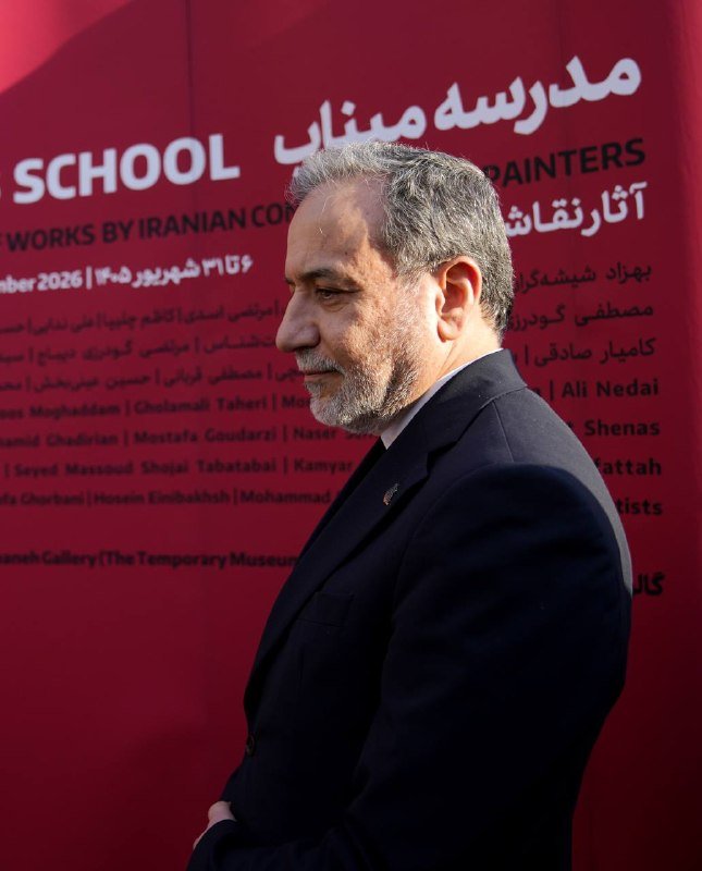 A gray-bearded man in a dark suit stands in profile before a red backdrop displaying event text including "SCHOOL," "PAINTERS," and "WORKS BY IRANIAN CO..."