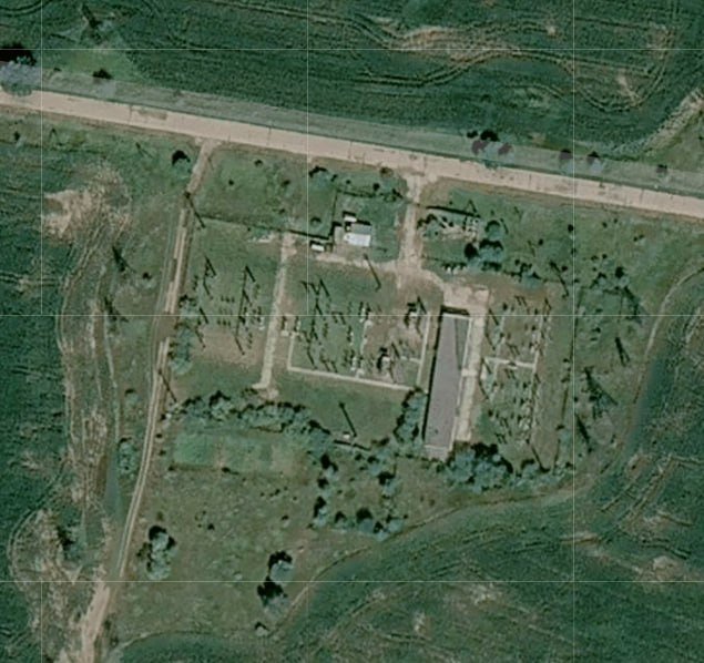 An aerial satellite image shows a clearing with rows of trees, small structures, and a long rectangular building surrounded by green terrain.