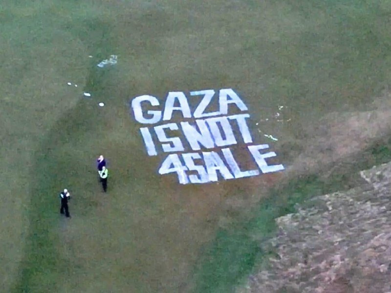 Large white letters spelling "GAZA IS NOT 4 SALE" are arranged on a grassy hillside, with two small figures standing nearby on the grass.