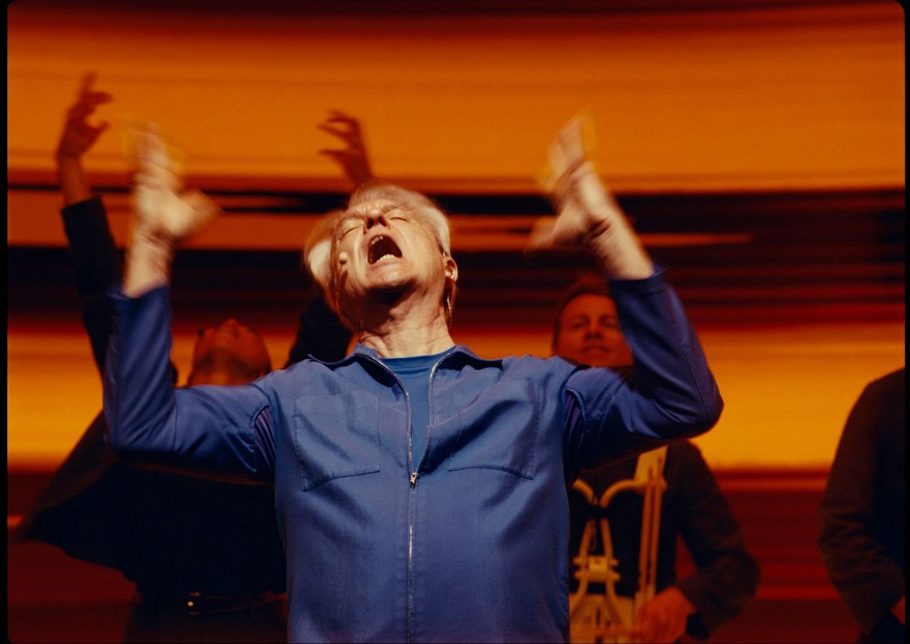 A performer in a blue zip-up shirt sings passionately with mouth wide open and arms raised, backed by musicians in a warmly lit orange-hued setting.