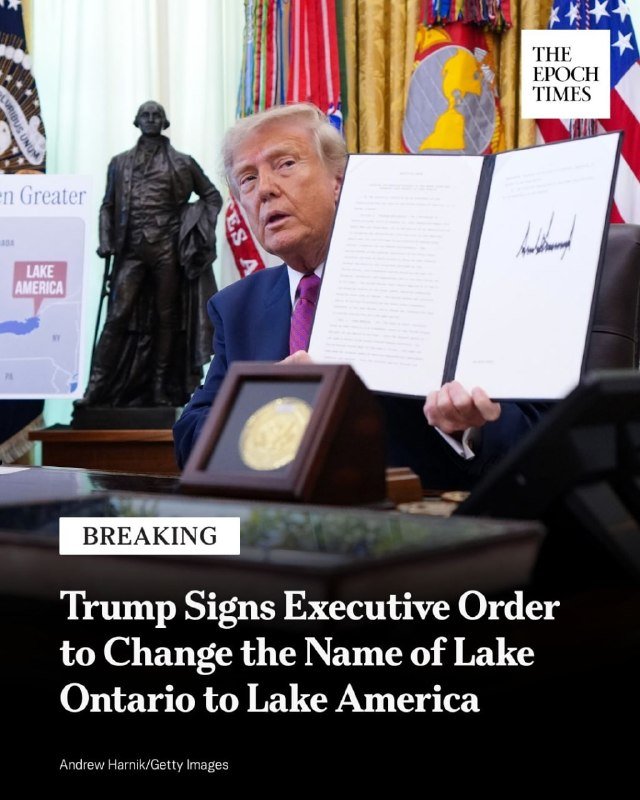 A man in a dark suit and purple tie holds up a signed document displaying his signature, with American flags and a bronze statue visible in the background.