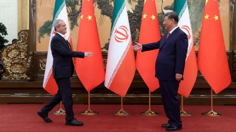 Two men in dark suits reach out to shake hands on a red carpet, standing before alternating Chinese and Iranian flags.