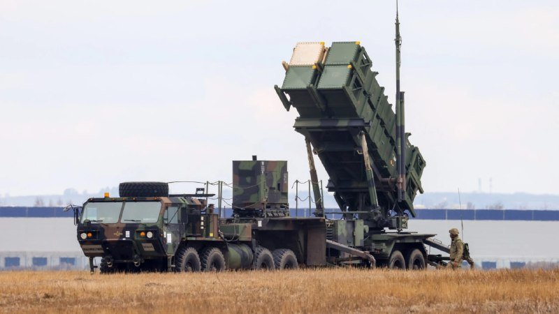 A camouflaged military truck carries a raised, angled missile launcher system in a grassy field, with a soldier in uniform walking nearby.