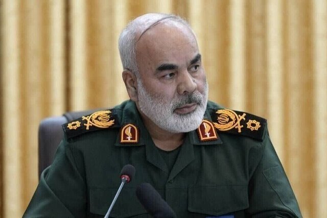 A bearded man in a green military uniform with gold epaulets sits before a microphone against a beige curtain backdrop.