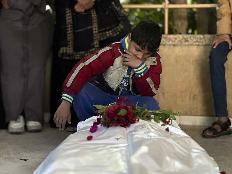 A boy in a red and white jacket crouches beside a white-shrouded body covered in red flowers, wiping his face while other people stand nearby.