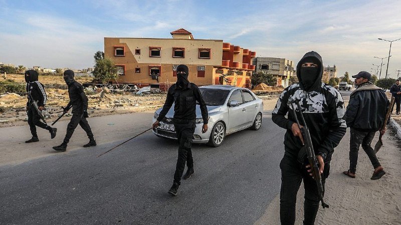Masked, armed individuals in black clothing walk along a paved road past a silver sedan, with orange and tan buildings visible in the background under a partly cloudy sky.
