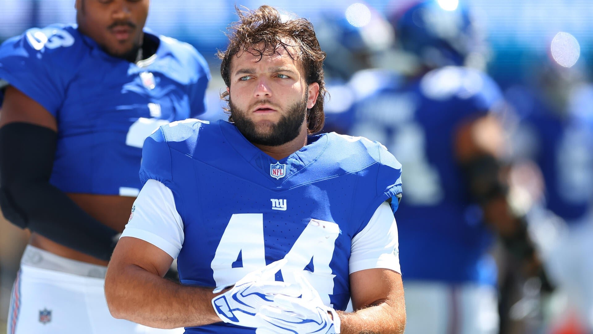 A bearded football player wearing a blue New York Giants jersey stands on the sidelines, holding his helmet while other players are visible in the background.