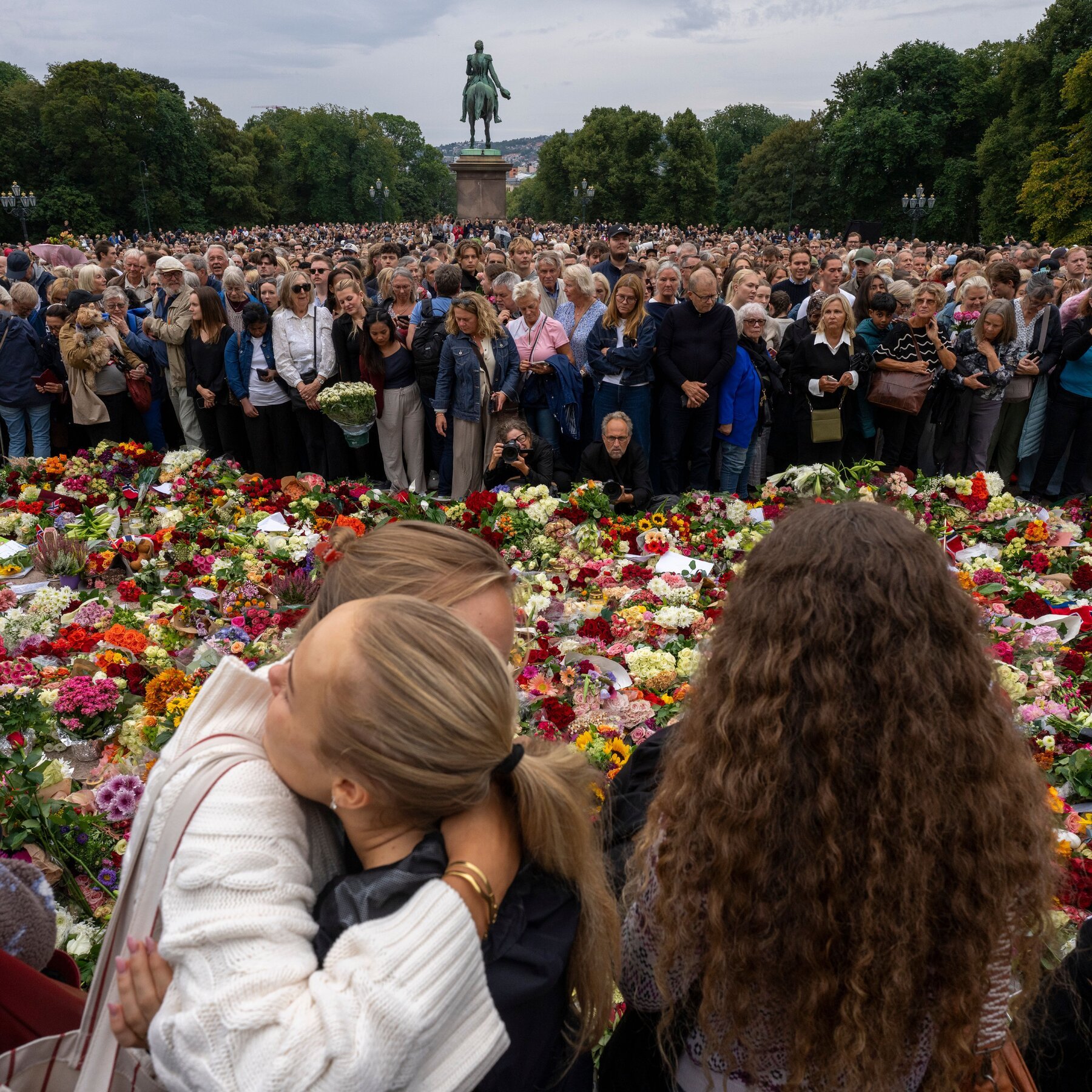 A large crowd gathers solemnly in a park before a mounted equestrian statue, with an extensive carpet of colorful flower bouquets laid out in the foreground.
