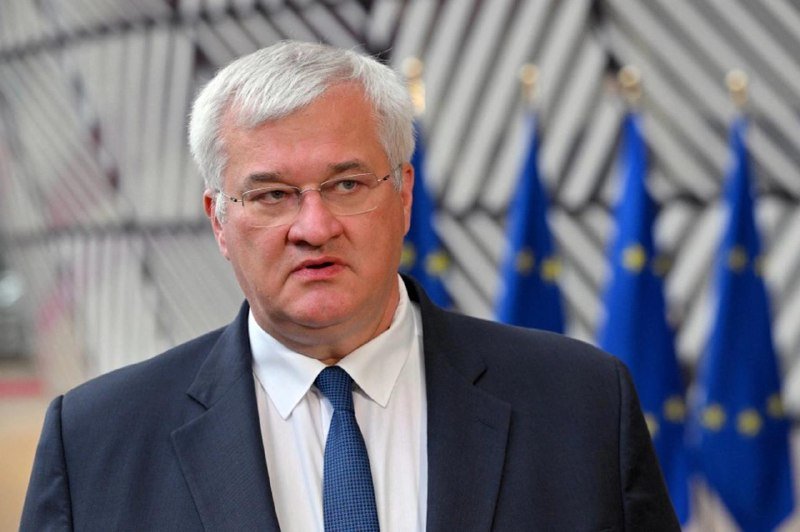 A gray-haired man in a dark suit, white shirt, and blue tie speaks while standing before a row of European Union flags.