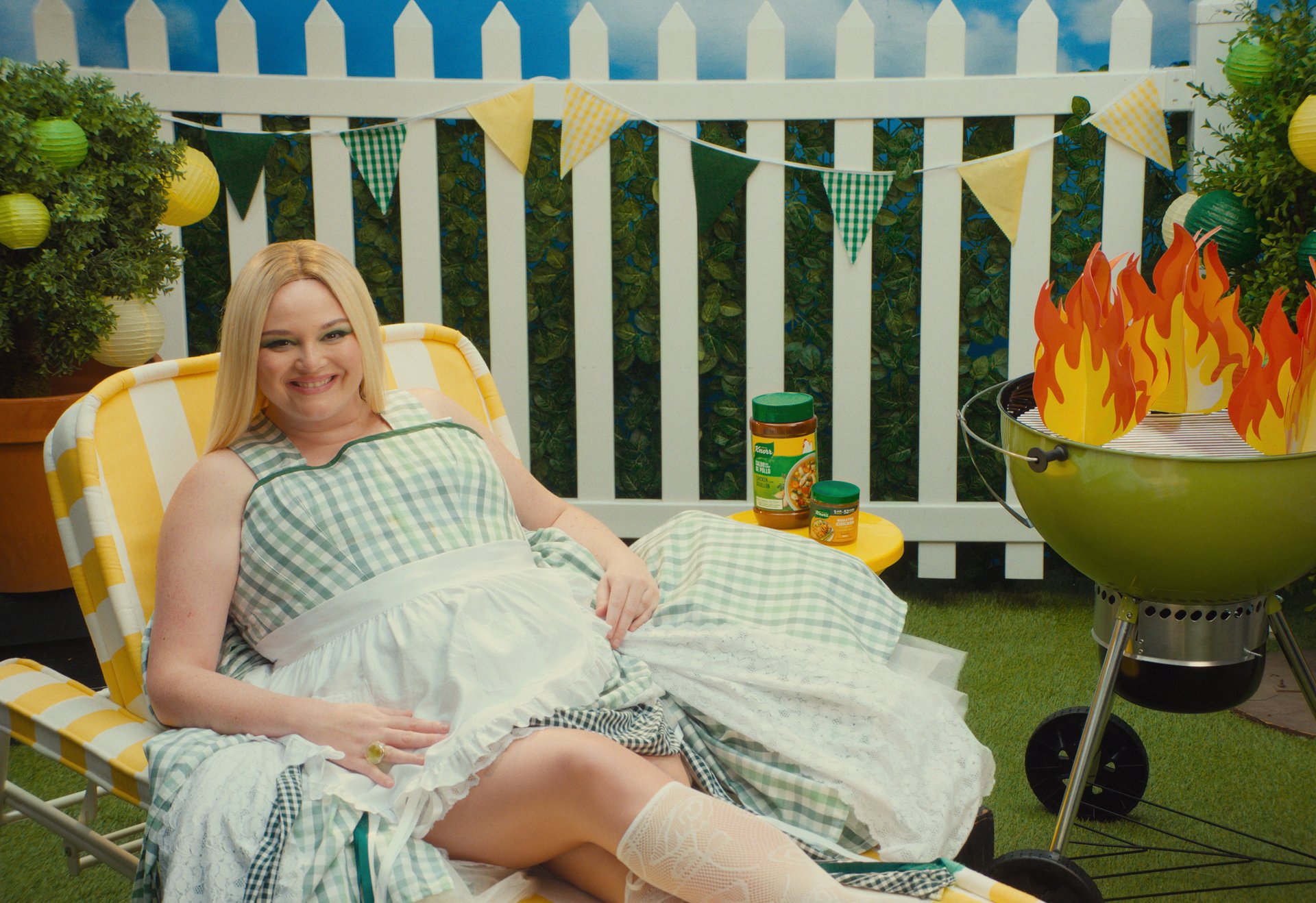 A blonde woman in a green gingham dress sits in a striped lawn chair beside a green grill decorated with paper flames, set against a white picket fence with bunting.