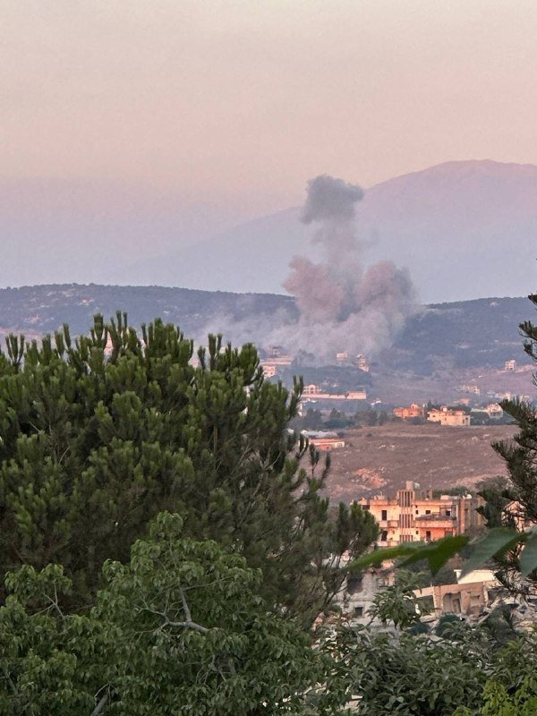 Plumes of gray smoke rise over a hillside settlement nestled between forested mountains, with green foliage and light-colored buildings visible in the foreground and valley below.
