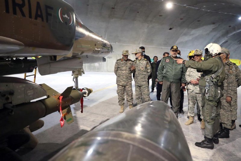 Military personnel in camouflage uniforms and flight gear stand beside an aircraft marked "IRIAF" inside an illuminated hangar, with ordnance visible beneath the plane's wing.