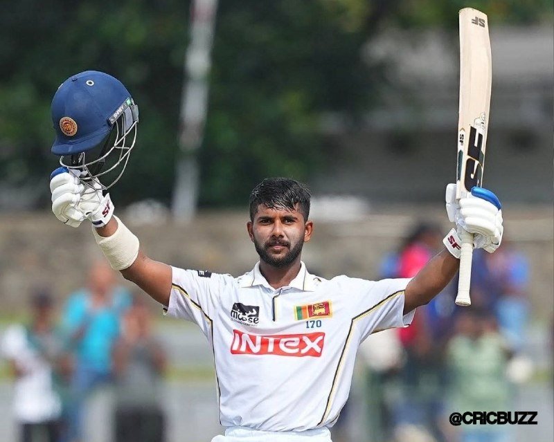 A bearded cricket player in a white "Intex" jersey raises his helmet in one hand and a bat in the other, celebrating on the field with a blurred crowd in the background.