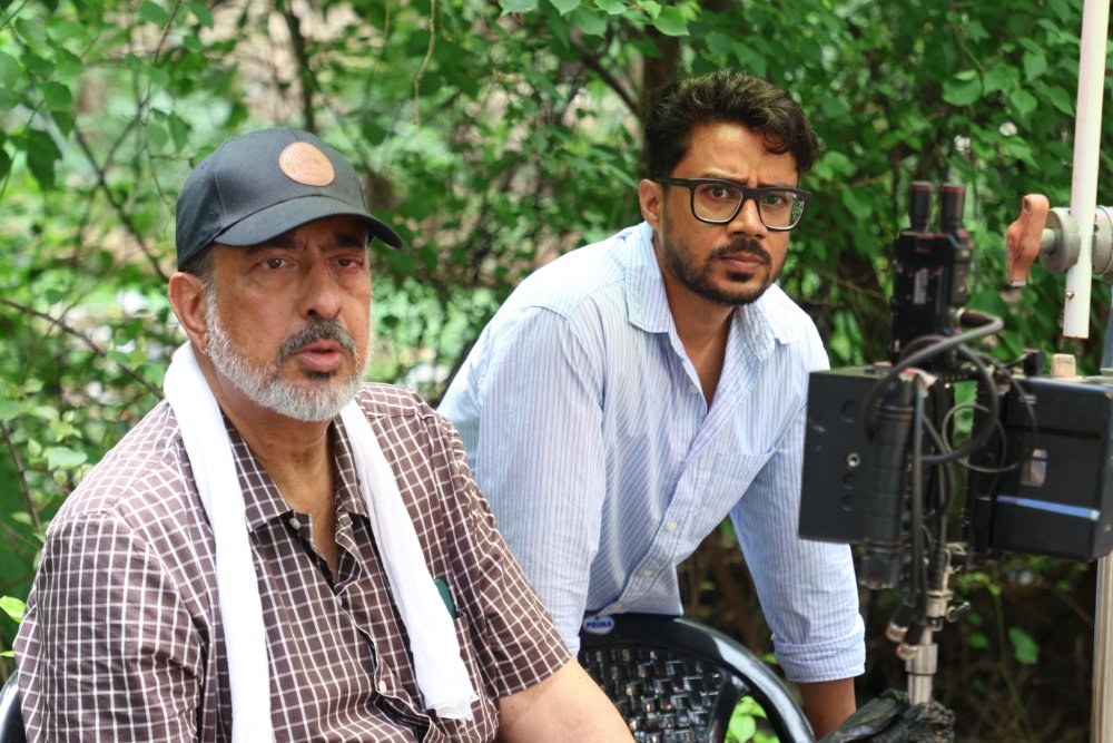 Two men are positioned on an outdoor film set, with one wearing a cap and checkered shirt seated while the other, in glasses, looks toward a professional camera rig against a leafy backdrop.