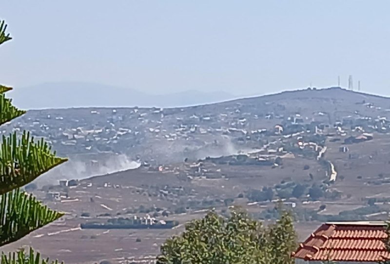 A plume of white smoke rises from a hillside among scattered buildings in a hazy, mountainous landscape, with tree foliage and a tiled roof in the foreground.