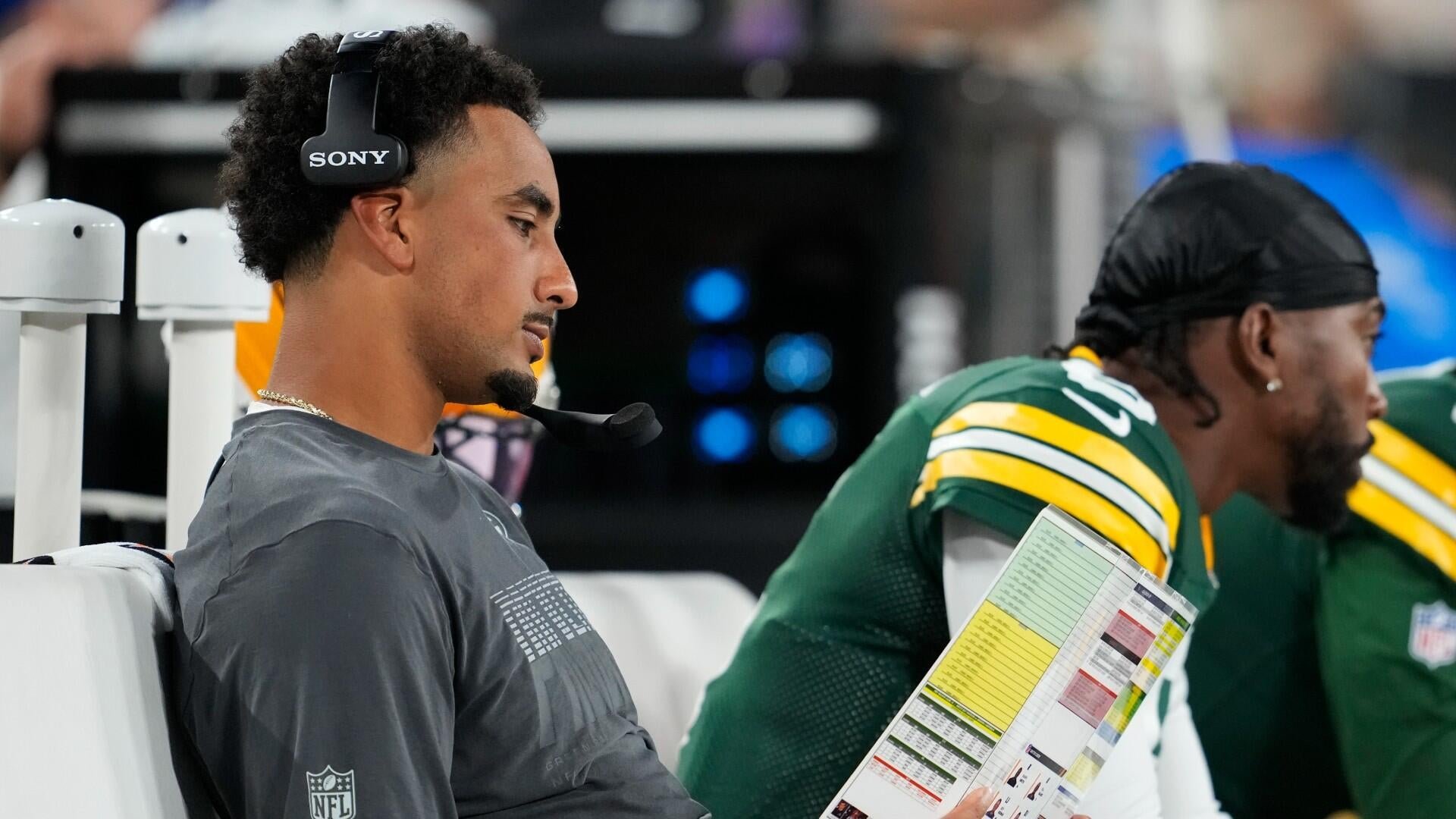 A man wearing a Sony headset and gray NFL shirt sits on a bench beside a player in a green and yellow jersey holding a play sheet.