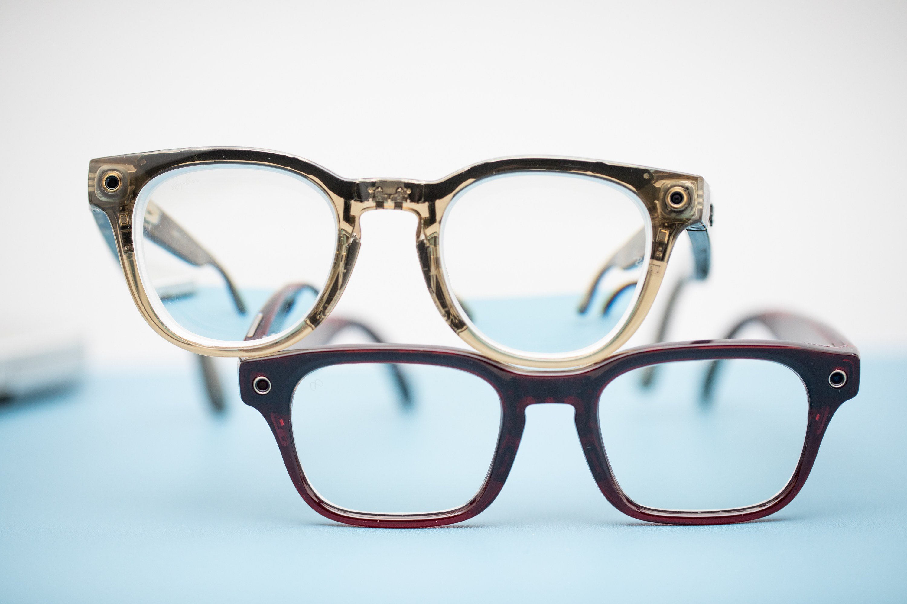 Two pairs of smart glasses—one transparent with visible internal components, the other dark red—are displayed on a light blue surface.