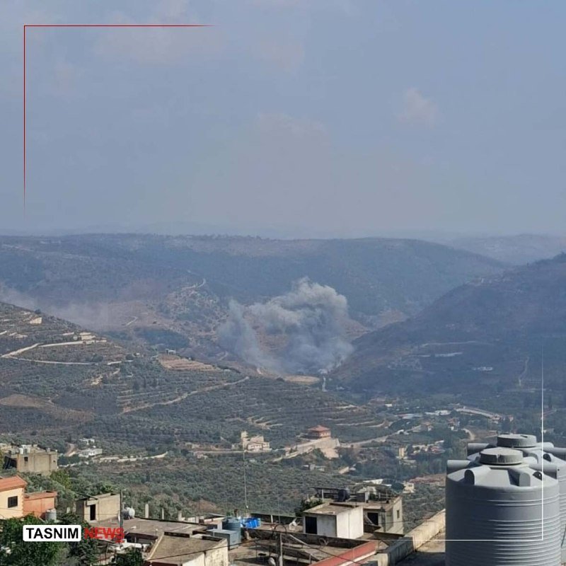 A plume of gray smoke rises from a hilly, terraced landscape dotted with buildings and water tanks, with the "TASNIM NEWS" logo in the corner.