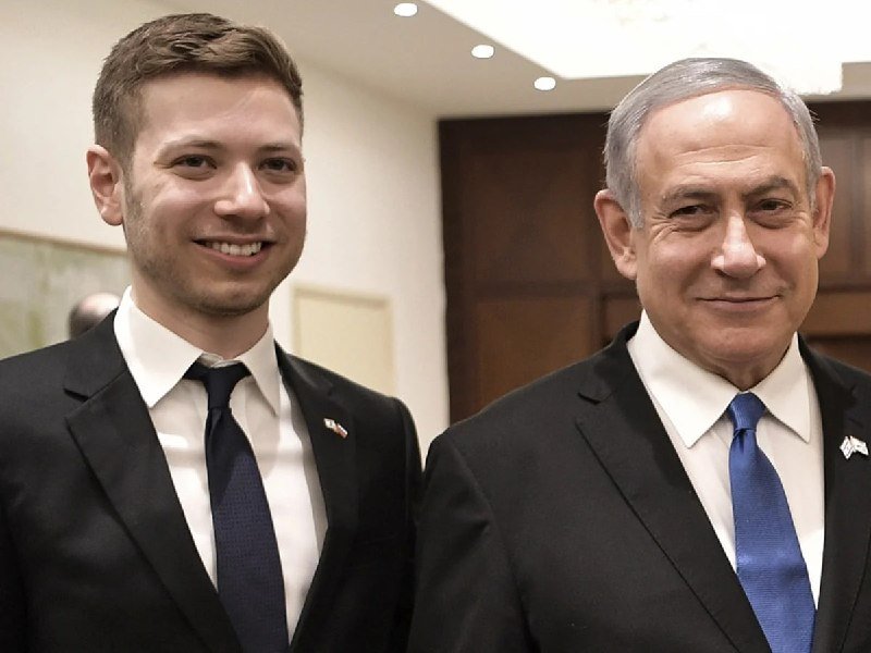 Two men in dark suits and ties, both wearing small lapel pins, stand side by side smiling indoors near a wooden door.