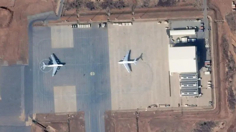 Aerial view of two large aircraft parked on an asphalt tarmac adjacent to a white-roofed hangar and storage area surrounded by bare earth.