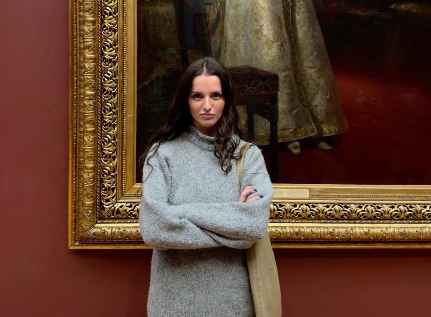 A woman with long dark hair, wearing a gray sweater, stands with arms crossed in front of a large, ornately framed painting displayed on a red wall.