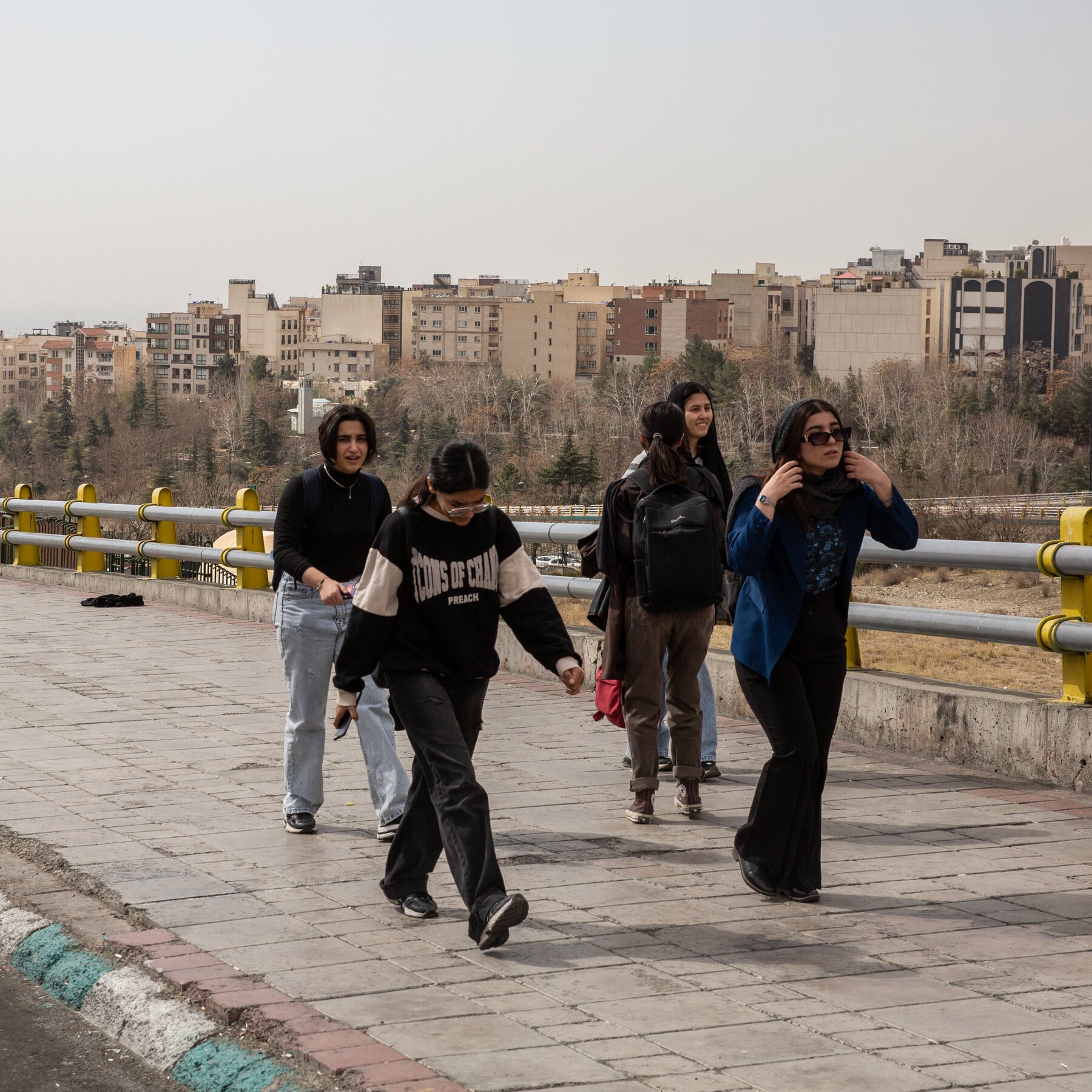 A group of young women walk across a paved bridge with yellow railings, wearing casual clothing and backpacks, with a hazy city skyline and trees in the background.