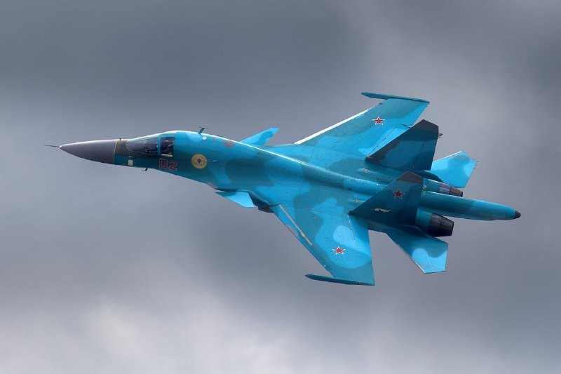 A blue camouflaged fighter jet with red star markings and the number "02" banks sharply through a cloudy gray sky.