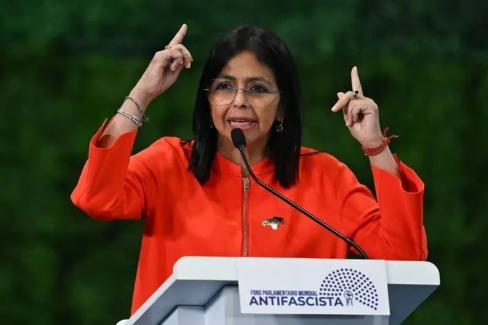 A woman with glasses in an orange jacket speaks at a podium with both hands raised, gesturing with her index fingers pointing upward.