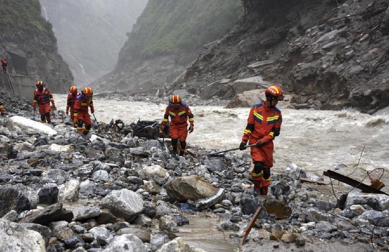 Rescue workers in orange uniforms and helmets navigate a rocky riverbank beside rushing water, carrying equipment through a steep, debris-strewn mountain gorge.