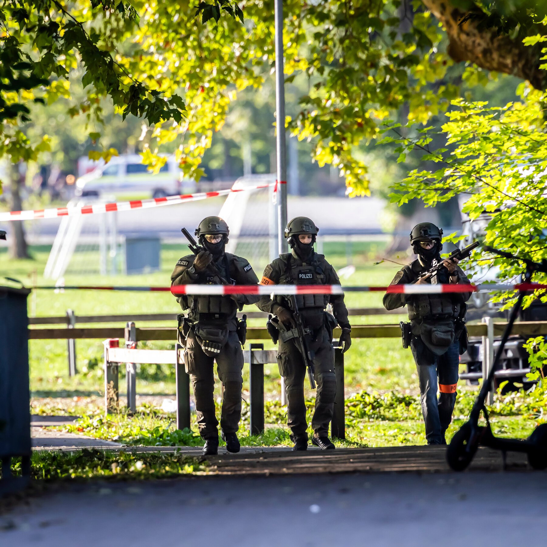 Three armed officers in tactical gear marked "POLIZEI" stand behind red-and-white police tape in a tree-lined park setting.