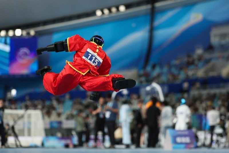 A competitor in a red snowsuit and goggles, wearing bib number 115, performs a mid-air trick inside an arena.