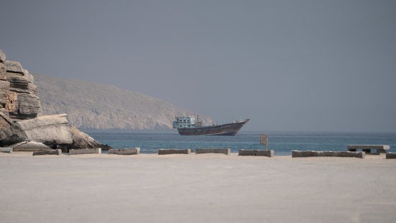 A large cargo ship floats on calm blue waters near a rocky coastline, with a sandy shore and concrete barriers visible in the foreground.