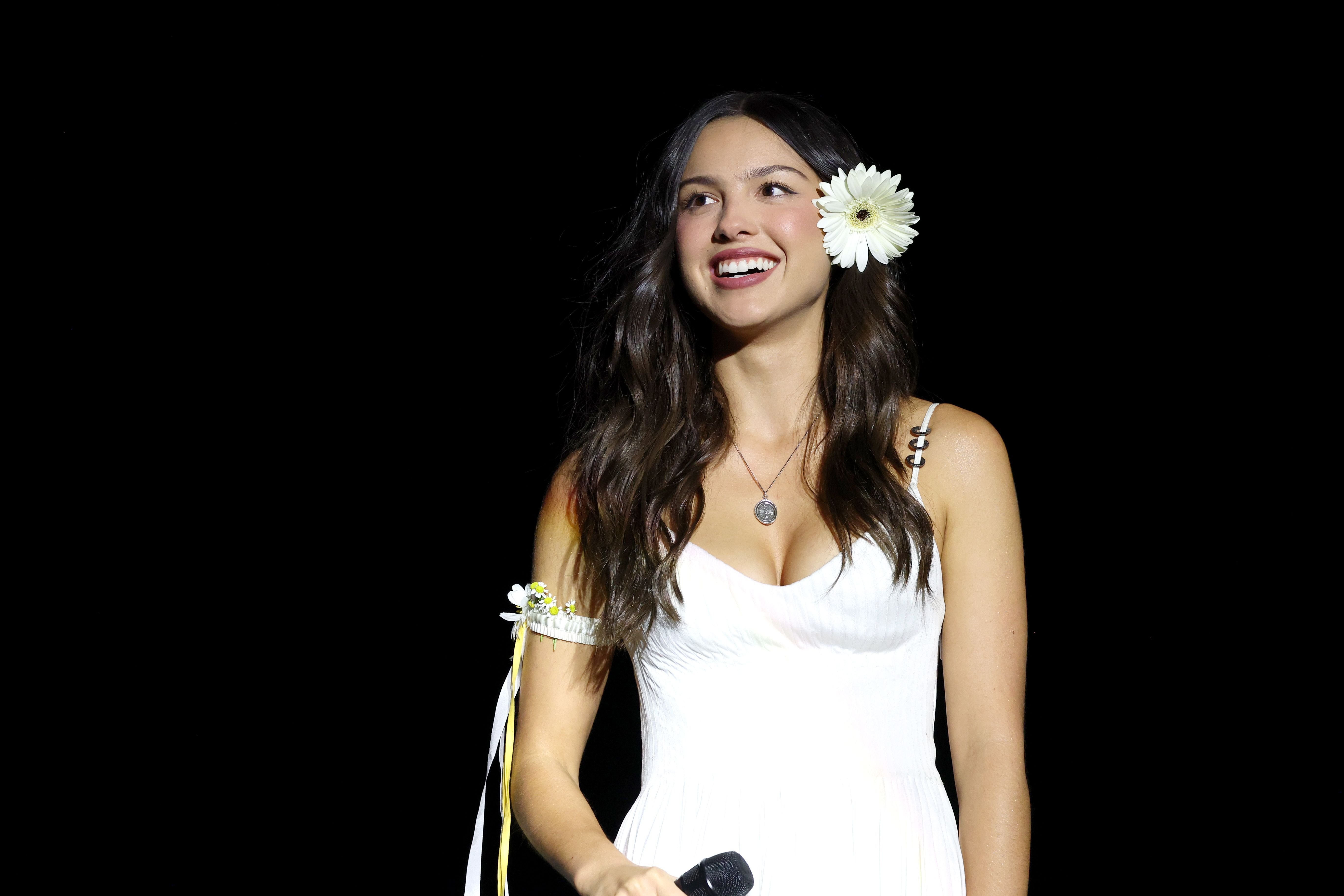 A smiling woman with long dark hair, wearing a white dress and a white flower behind her ear, holds a microphone against a dark background.