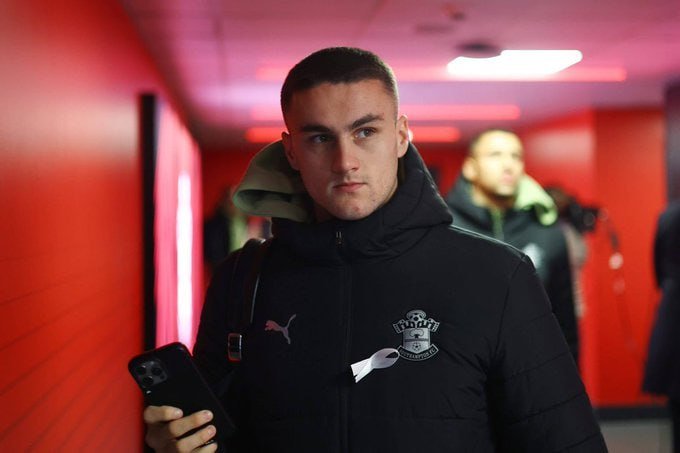 A young man wearing a black Southport FC jacket walks through a red-lit corridor holding a phone, with another person visible behind him.