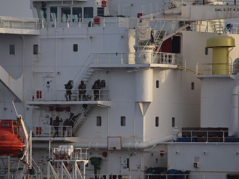 Armed personnel in dark tactical gear ascend a white staircase on the multi-deck superstructure of a large white vessel, with an orange lifeboat visible at lower left.