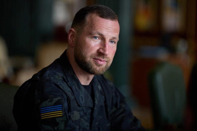 A bearded man in a dark camouflage uniform with a blue and yellow striped patch sits indoors, looking toward the camera.