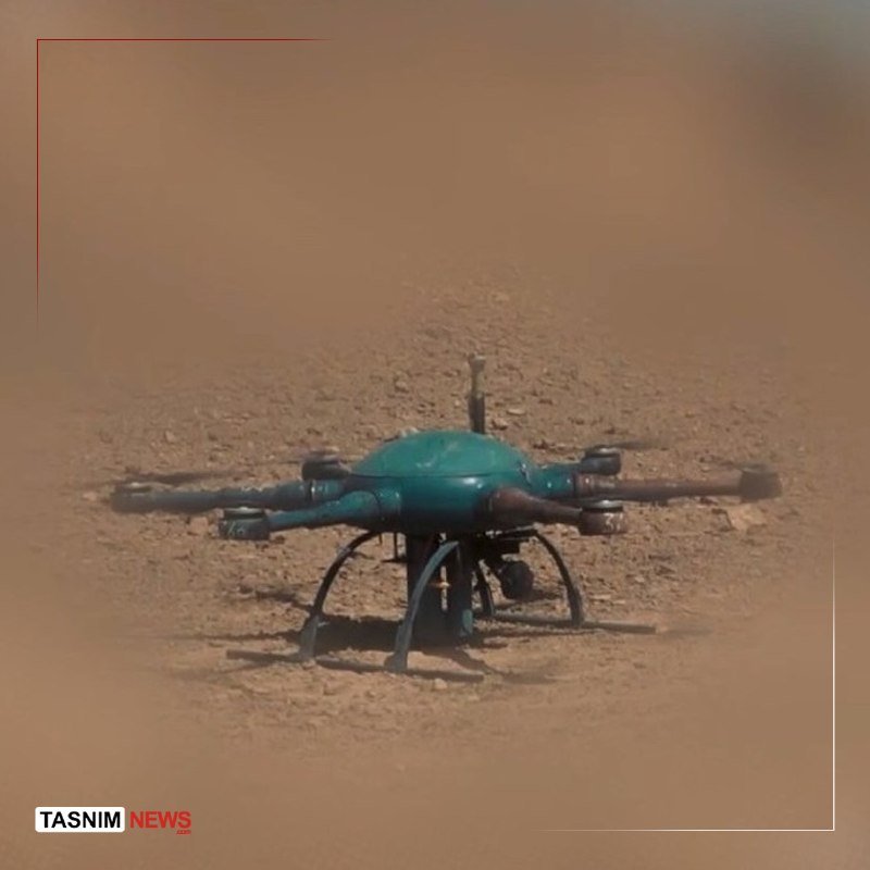 A teal quadcopter drone rests on a sandy, dirt surface, with a "TASNIM NEWS" logo visible in the bottom left corner.