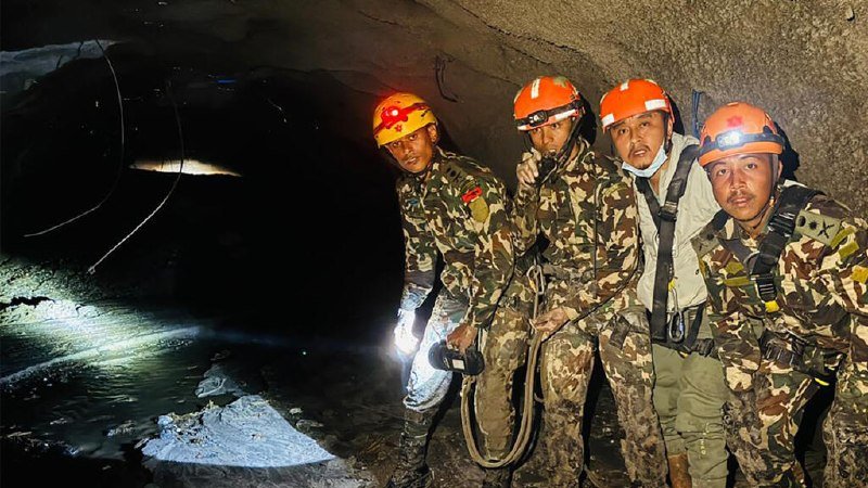 Four men in camouflage uniforms and orange helmets with headlamps stand inside a dark, rocky cave beside a shallow stream.