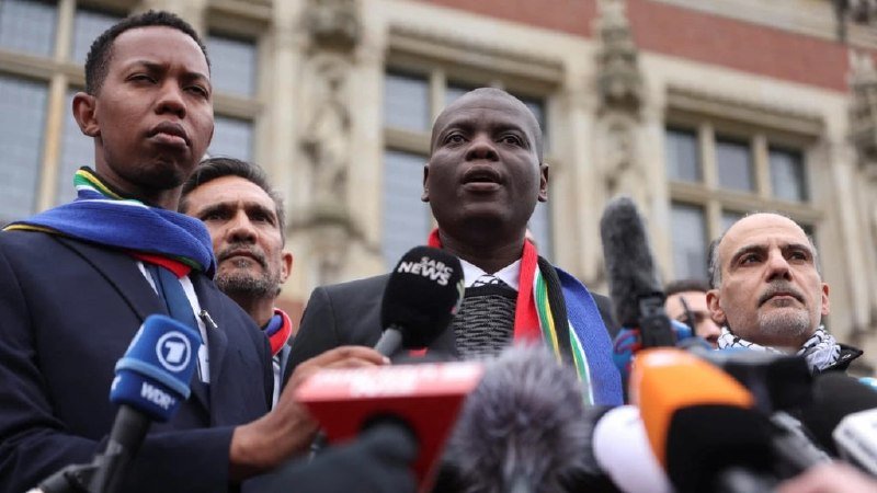 Four men stand outside an ornate stone building while a man wearing a multicolored scarf speaks into a cluster of media microphones.