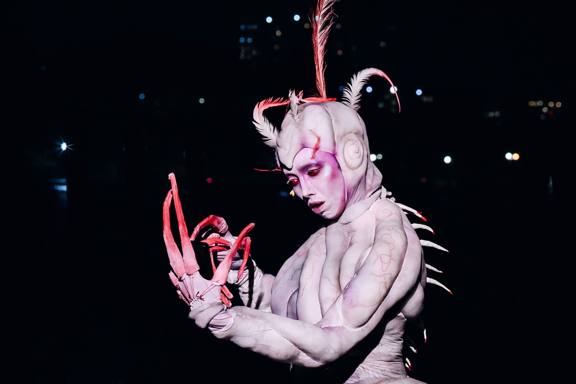 A performer in pale body paint with vein-like markings, feathered headpiece, and spiked spine holds large red claw-like hands against a dark background of blurred city lights.