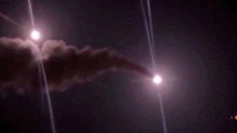A bright projectile trails thick smoke and fire as it streaks across the night sky, illuminated by two bright light sources.
