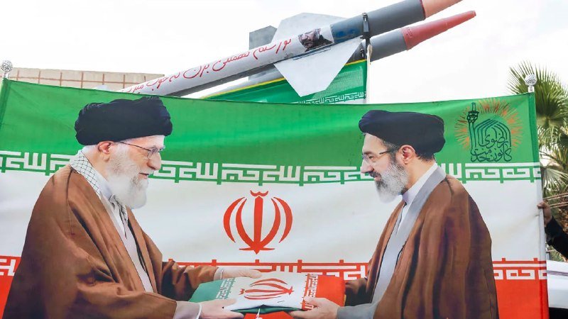 Two bearded men wearing black turbans and clerical robes stand before an Iranian flag and banner, jointly holding a small flag as a model missile is displayed behind them.
